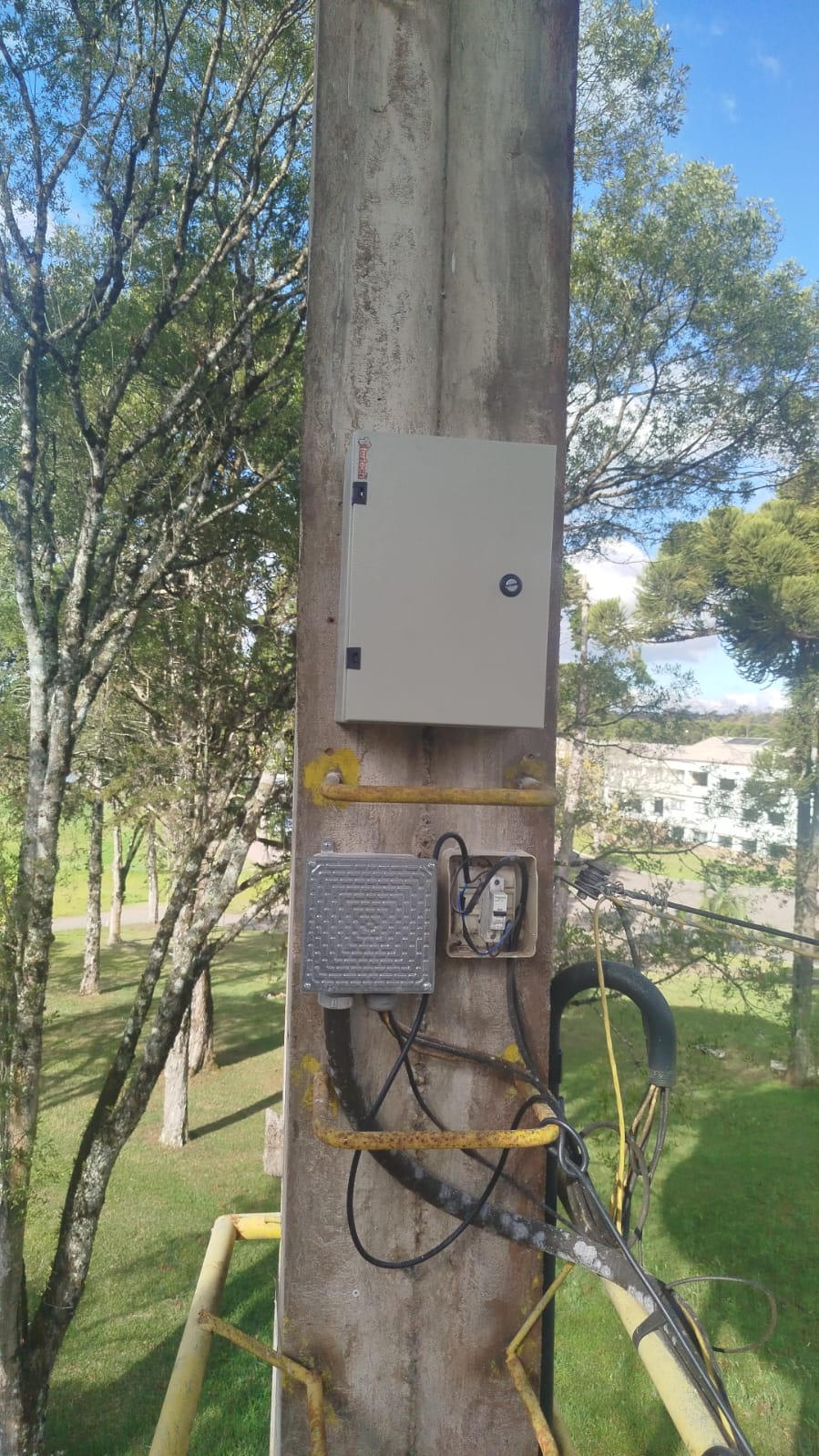 caixa elétrica metálica e caixas de passagem com fios instaladas num poste de concreto, visto de cima com árvo