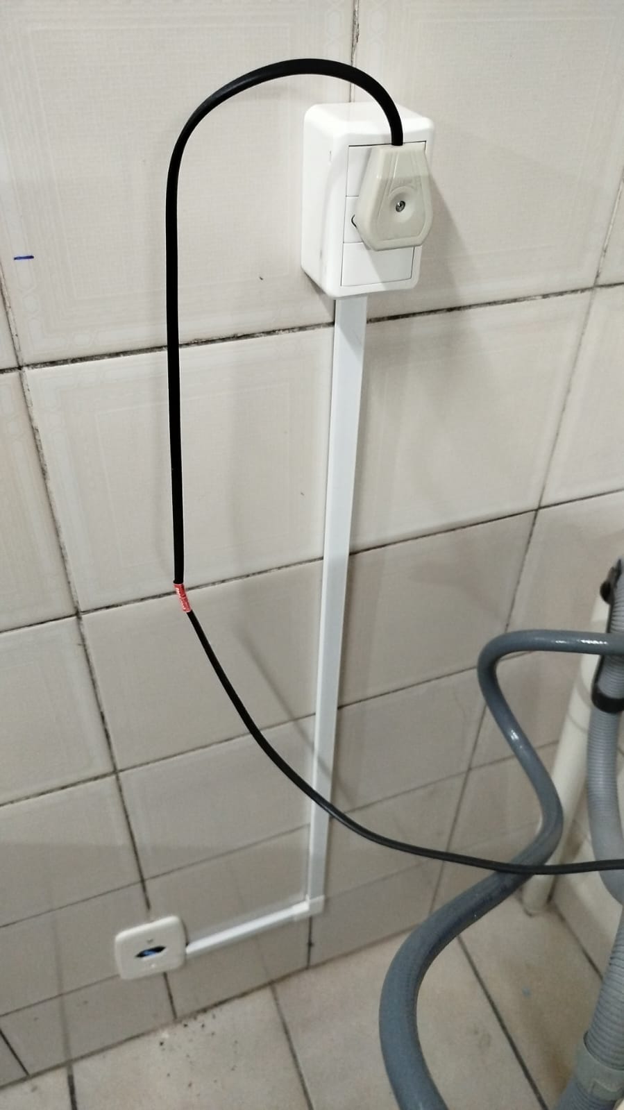 tomada e canaleta elétrica instaladas em parede de azulejos, com cabo preto e mangueira cinza de máquina de la