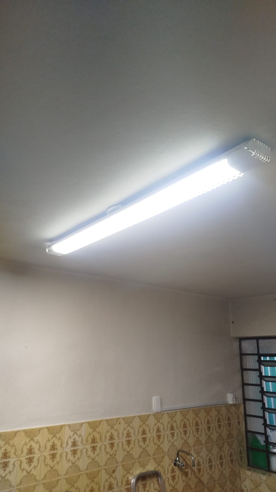 luminária LED de tubo duplo instalada no teto de uma cozinha com azulejos amarelos e pia ao fundo — texto: 