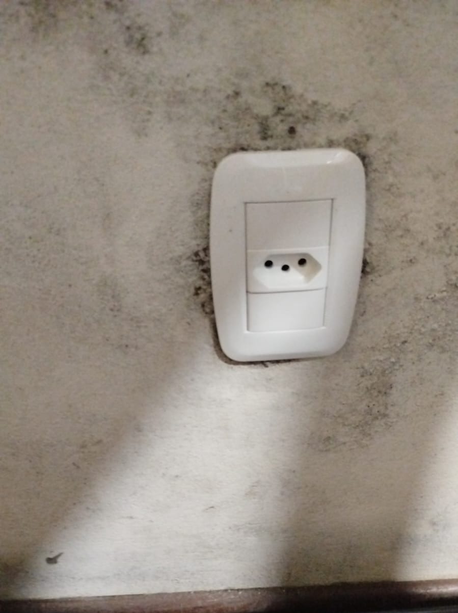 tomada elétrica branca de dois pinos instalada em parede manchada de mofo, com rodapé de madeira e piso escuro